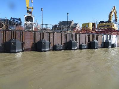 Metal baffle along a riverfront. Construction and excavation equipment  on a walkway above the river and behind a railing. The river water is brown.