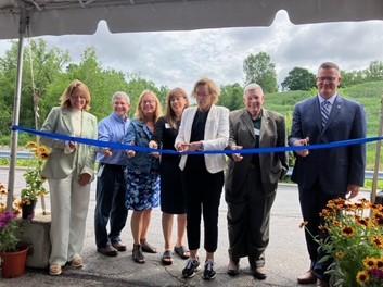 Seven people reach out with scissors to cut a blue ribbon in a ceremony outside.