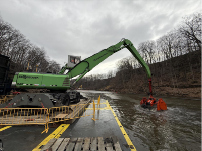Green silt barge dredges sediment from the Cuyahoga Gorge dam pool on an cloudy spring day.