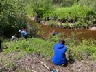 Six people along the banks of a stream. 5 of them are hunched over examining things on the ground. A sixth person is squatting in the forefront, his back to us, looking at the water.