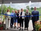 Seven people reach out with scissors to cut a blue ribbon in a ceremony outside.