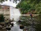 A person stands in a body of water near a flowing dam to collect a water quality sample.